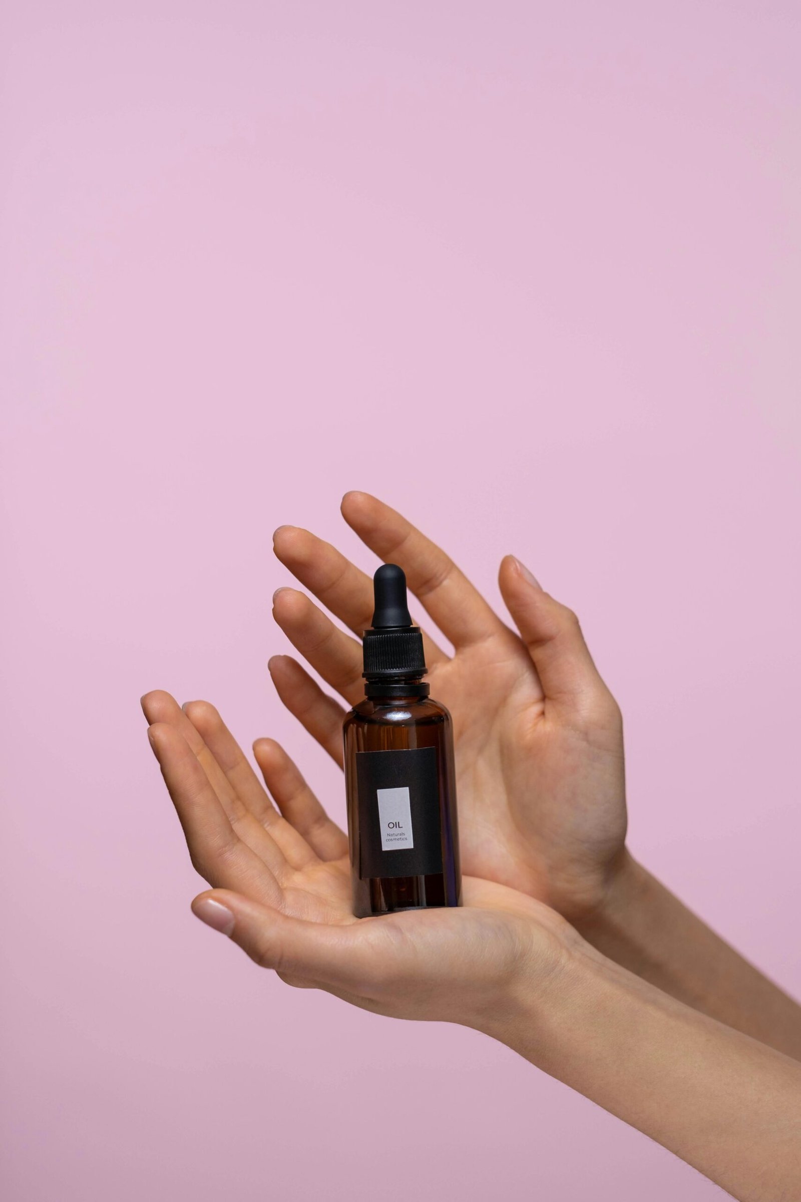 Close-up of a brown dropper bottle held by hands against a pink background, representing natural skincare.